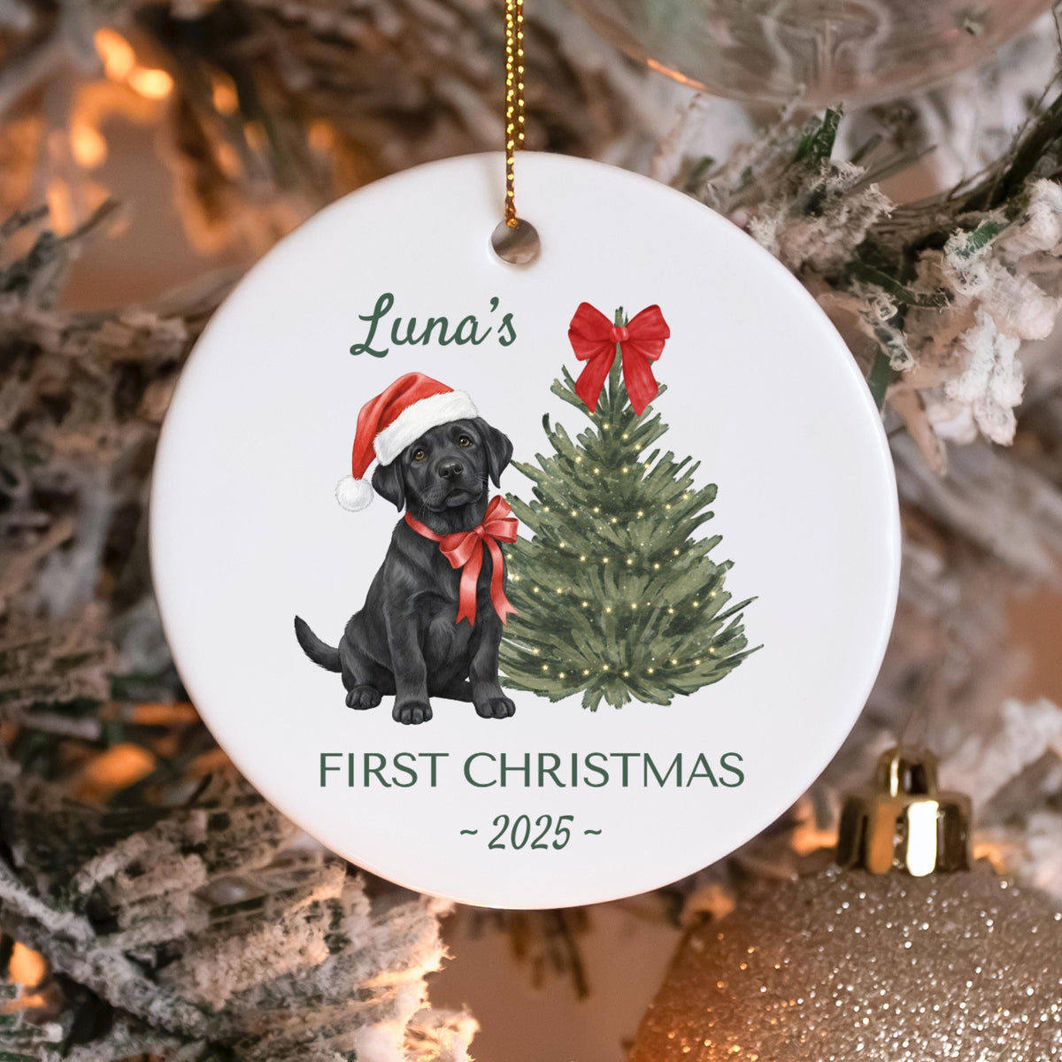 White ceramic Christmas ornament featuring a black Labrador puppy wearing a Santa hat and red bow. A green Christmas tree with lights and the text "Luna's First Christmas - 2025" are also displayed.