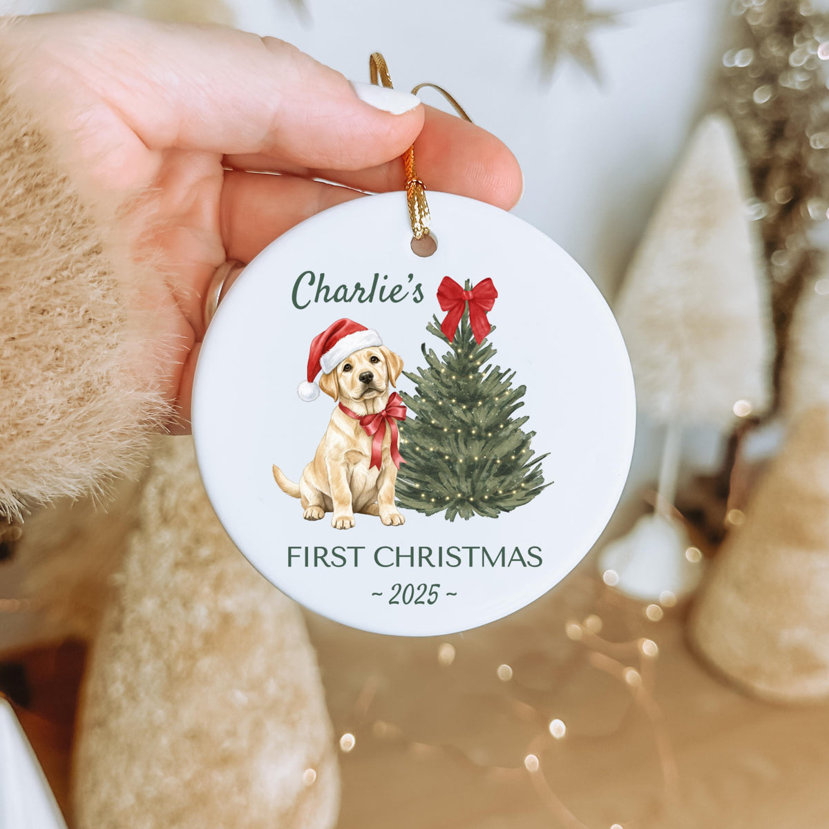 A round, white Christmas ornament featuring a watercolor illustration of a yellow Labrador puppy wearing a Santa hat and a red bow. The text "Charlie's FIRST CHRISTMAS - 2025 -" is printed on the ornament.