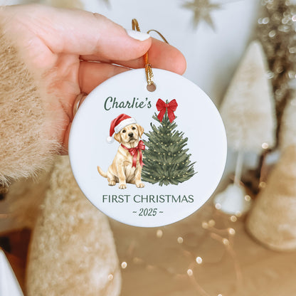 A round, white Christmas ornament featuring a watercolor illustration of a yellow Labrador puppy wearing a Santa hat and a red bow. The text "Charlie's FIRST CHRISTMAS - 2025 -" is printed on the ornament.