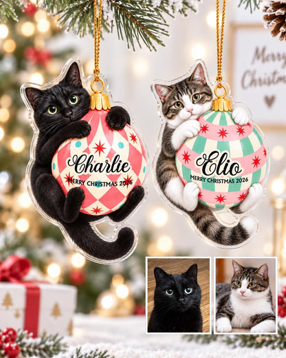 Two flat acrylic Christmas ornaments hanging by gold strings. One features a black cat hugging a pink and white checkered bauble labeled Charlie, the other a tabby cat hugging a pink and green striped bauble labeled Elio. Both say Merry Christmas 2026.