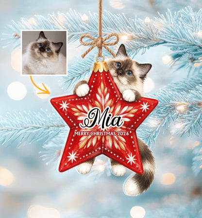 A star-shaped Christmas ornament featuring a fluffy cat with dark ears and a tail, holding onto the red star. The ornament is printed with the name Mia and the text Merry Christmas 2026, suspended by a brown twine bow.