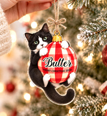 A flat, clear acrylic Christmas ornament shaped like a black and white tuxedo cat hugging a red and white checkered ball. The ball features the text Butter and MERRY CHRISTMAS 2026 in black script and sans-serif font, with a twine loop.