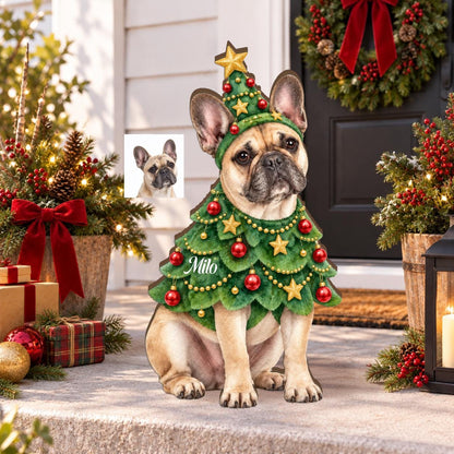 A tan French Bulldog wearing a green Christmas tree costume with red ornaments, gold stars, and a matching tree-shaped hat. The costume features the name Milo in white script. A small inset photo shows the dog without the costume.