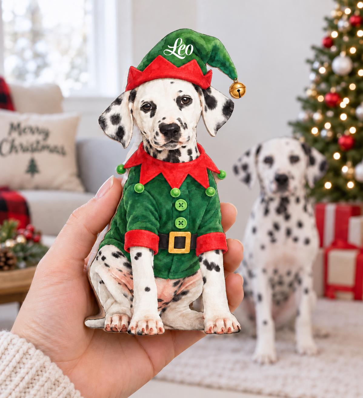 A flat ornament shaped like a Dalmatian dog wearing a green and red elf costume with a matching hat. The hat features the name Leo in white script and a small gold bell. The dog has black spots and is dressed in a green jacket with red trim, green buttons, and a black belt with a gold buckle.