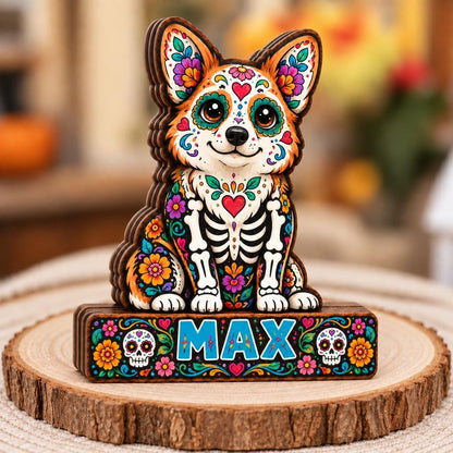 A wooden cutout of a corgi dog featuring colorful floral patterns and skeleton motifs in a Day of the Dead style. The base is a rectangular wooden block with the name MAX printed in blue, flanked by two white sugar skull illustrations.
