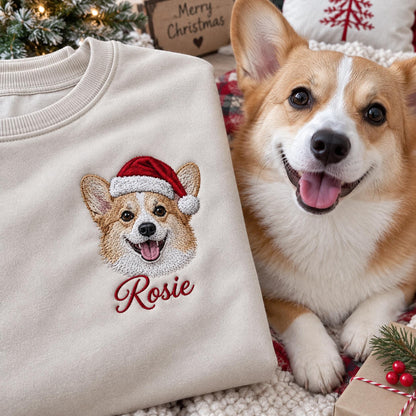 A beige crewneck sweatshirt featuring an embroidered design of a Corgi wearing a red and white Santa hat. Below the dog, the name Rosie is embroidered in red script. The sweatshirt has a ribbed collar and long sleeves.