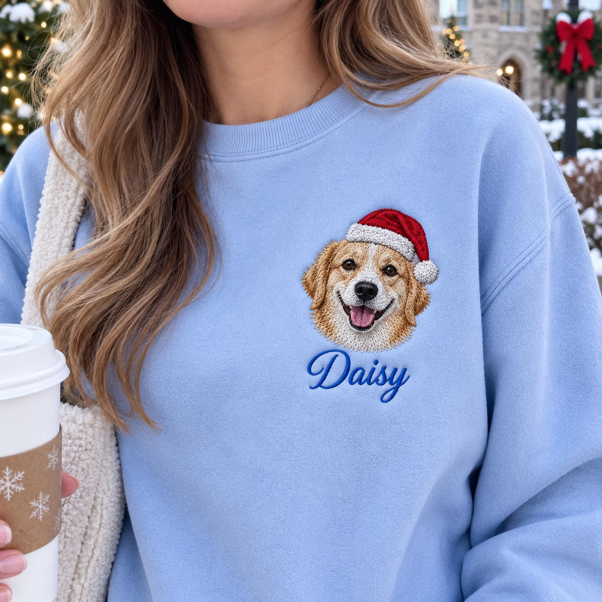 A light blue crewneck sweatshirt featuring an embroidered golden retriever wearing a red and white Santa hat. Below the dog, the name Daisy is embroidered in a dark blue cursive script. The fabric has a smooth, solid texture.