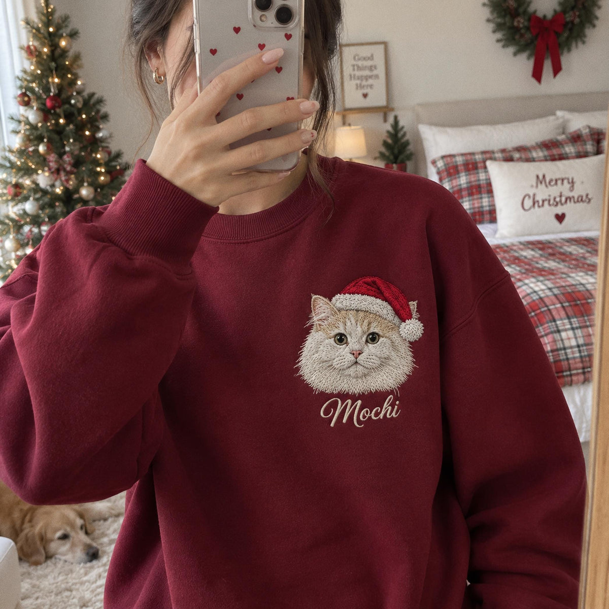 A dark red crewneck sweatshirt featuring an embroidered graphic of a fluffy cat wearing a red and white Santa hat. Below the cat, the name Mochi is embroidered in a white cursive script. The sweatshirt has long sleeves and ribbed cuffs.