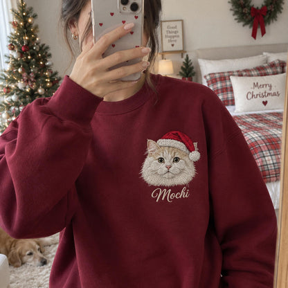 A dark red crewneck sweatshirt featuring an embroidered graphic of a fluffy cat wearing a red and white Santa hat. Below the cat, the name Mochi is embroidered in a white cursive script. The sweatshirt has long sleeves and ribbed cuffs.
