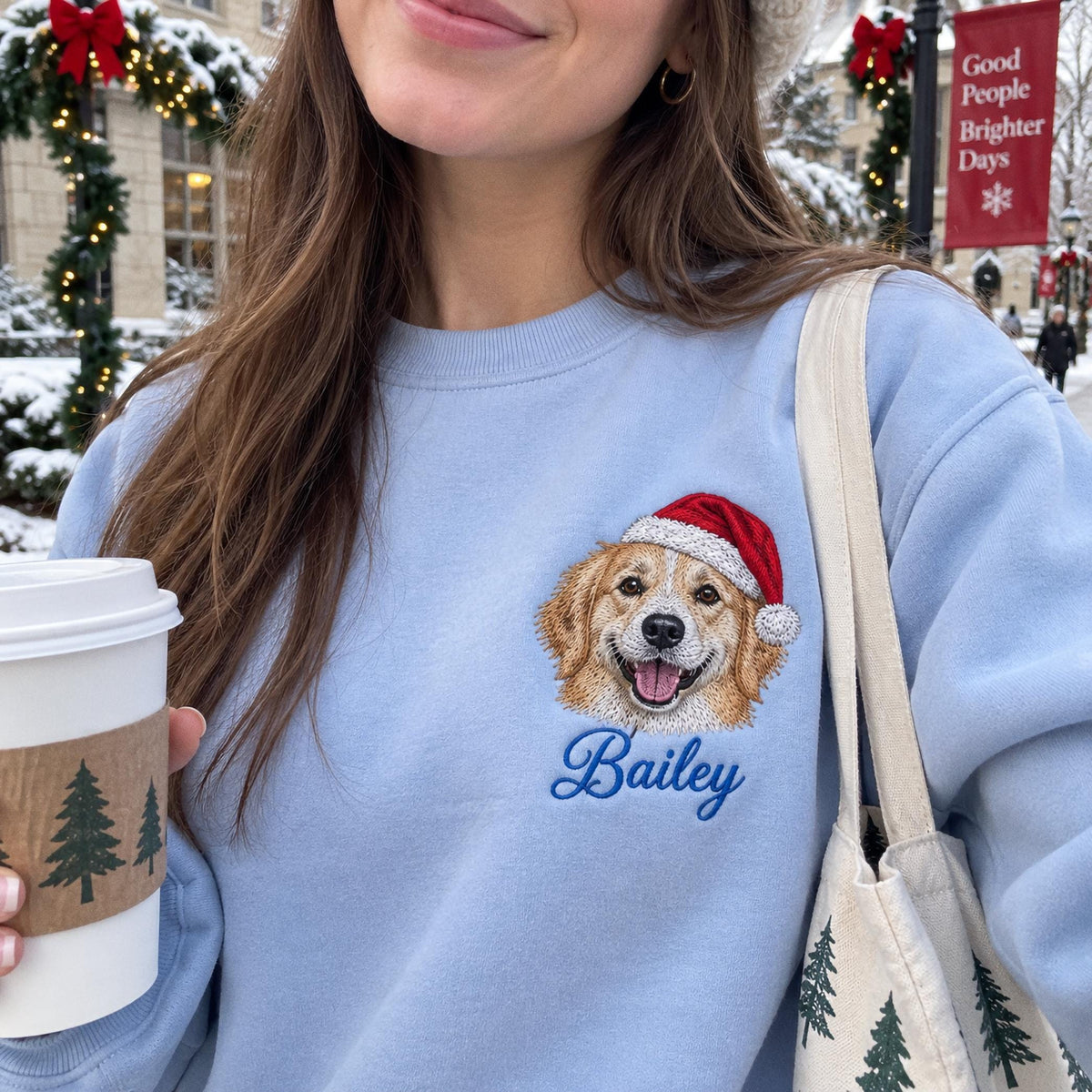 A light blue crewneck sweatshirt featuring an embroidered golden retriever wearing a red and white Santa hat. Below the dog, the name Bailey is embroidered in blue script. The sweatshirt is worn with a white tote bag with tree prints.