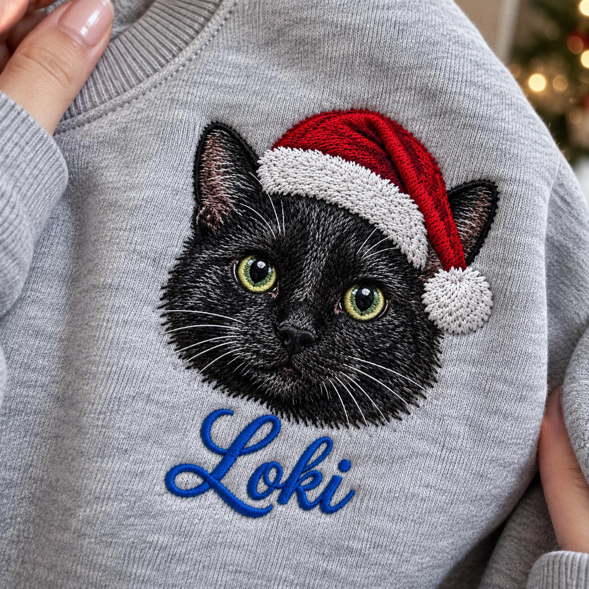 A light gray sweatshirt featuring an embroidered black cat wearing a red and white Santa hat. Below the cat, the name Loki is embroidered in blue cursive script. The design is centered on the chest of the garment.