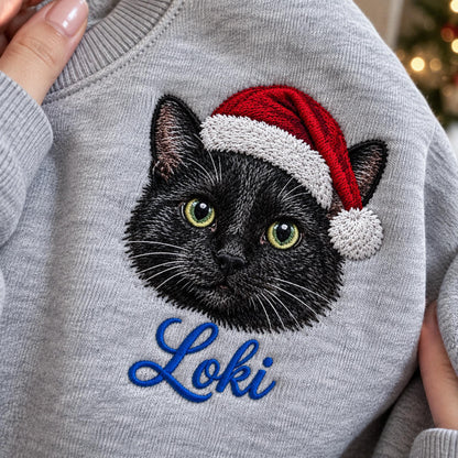 A light gray sweatshirt featuring an embroidered black cat wearing a red and white Santa hat. Below the cat, the name Loki is embroidered in blue cursive script. The design is centered on the chest of the garment.