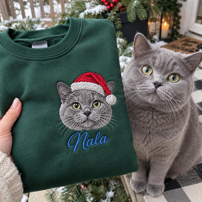 A dark green crewneck sweatshirt featuring an embroidered graphic of a grey cat wearing a red and white Santa hat. Below the cat, the name Nala is embroidered in blue script. A grey cat sits next to the sweatshirt, matching the design.