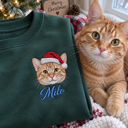 A dark green crewneck sweatshirt featuring an embroidered design of an orange tabby cat wearing a red and white Santa hat. Below the cat, the name Milo is embroidered in blue script. An orange tabby cat sits next to the sweatshirt.