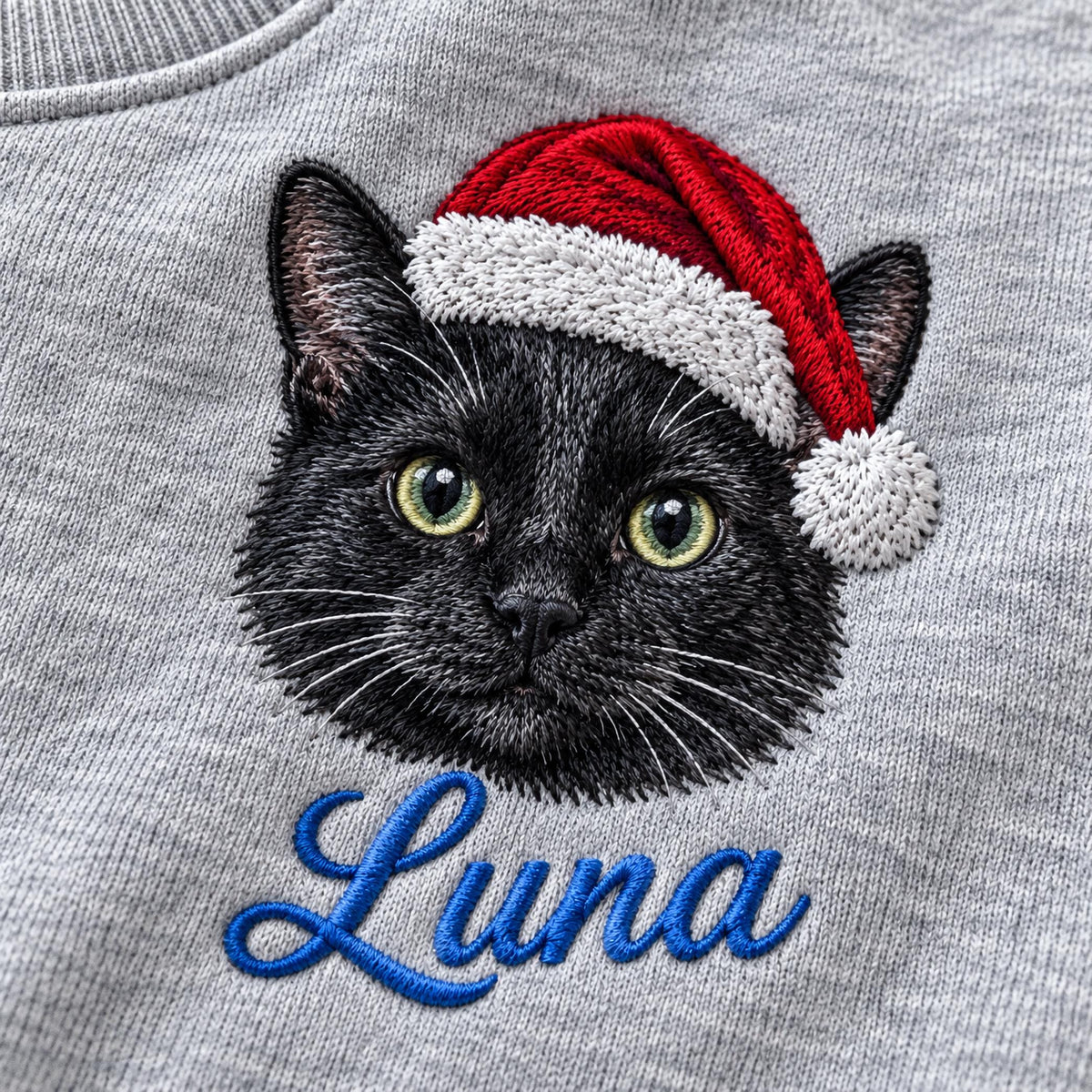 A heather gray sweatshirt featuring an embroidered black cat wearing a red and white Santa hat. Below the cat, the name Luna is embroidered in blue cursive script. The embroidery shows detailed fur texture and bright green eyes.