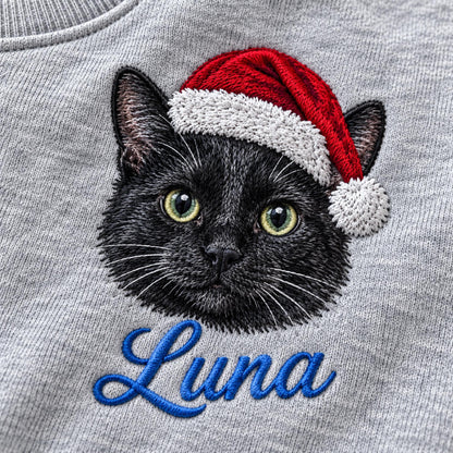 A heather gray sweatshirt featuring an embroidered black cat wearing a red and white Santa hat. Below the cat, the name Luna is embroidered in blue cursive script. The embroidery shows detailed fur texture and bright green eyes.