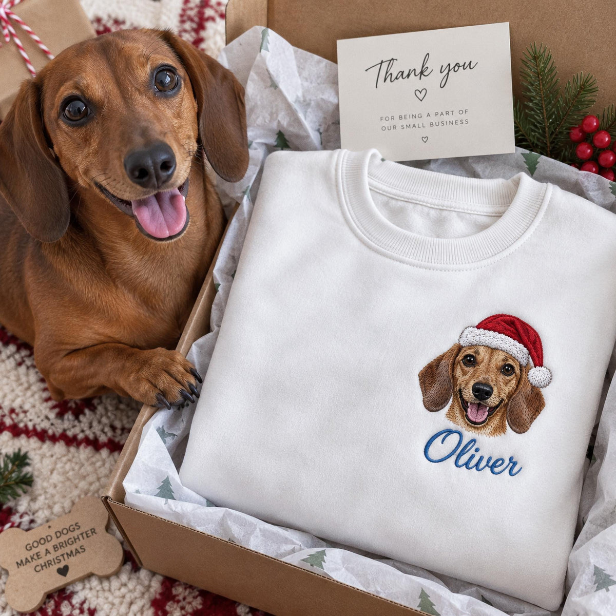 A white crewneck sweatshirt featuring an embroidered dachshund wearing a red and white Santa hat, with the name Oliver stitched in blue script below. The sweatshirt is packaged in a cardboard gift box with a thank you note.