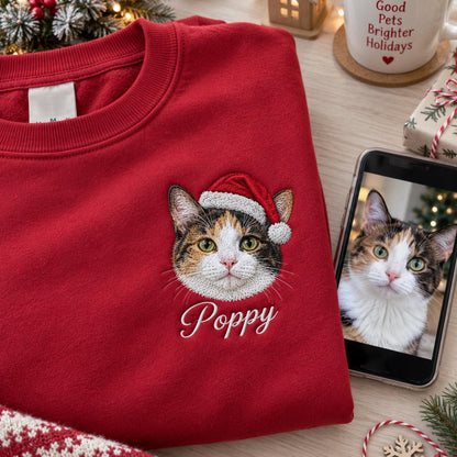 A red crewneck sweatshirt featuring an embroidered calico cat wearing a red and white Santa hat. Below the cat, the name Poppy is embroidered in white script. A smartphone displays a photo of the same cat next to the sweatshirt.