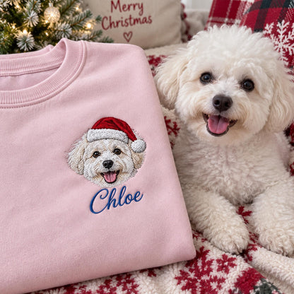 A light pink crewneck sweatshirt featuring an embroidered design of a white dog wearing a red and white Santa hat. Below the dog, the name Chloe is embroidered in blue script. The sweatshirt has a ribbed collar and long sleeves.