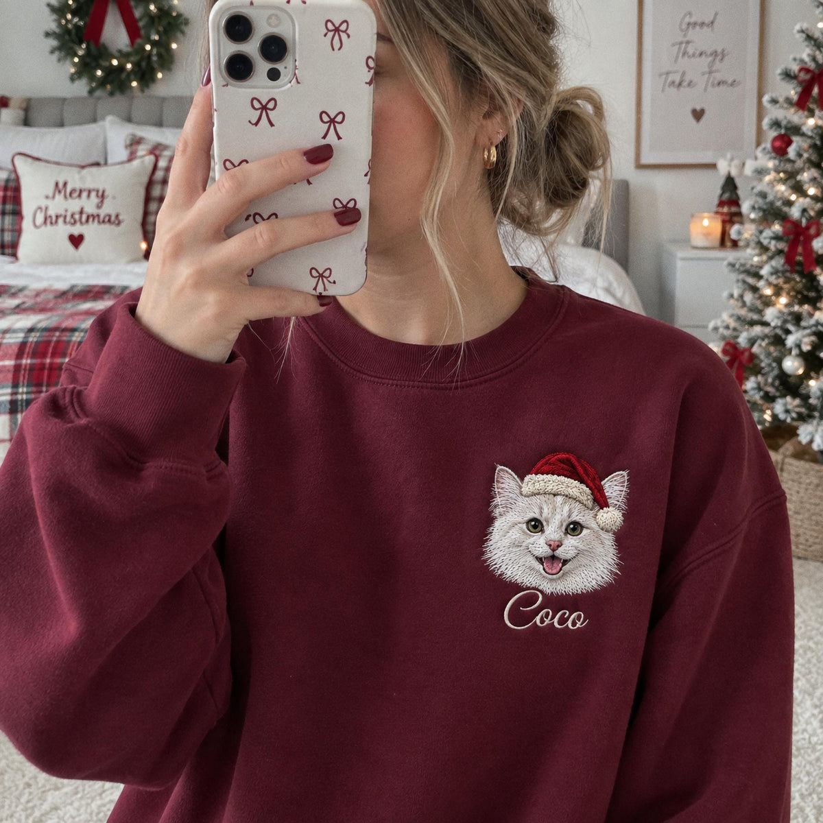 A maroon crewneck sweatshirt featuring an embroidered graphic of a white cat wearing a red and white Santa hat. Below the cat, the name Coco is embroidered in white script. The sweatshirt has long sleeves and a ribbed neckline.