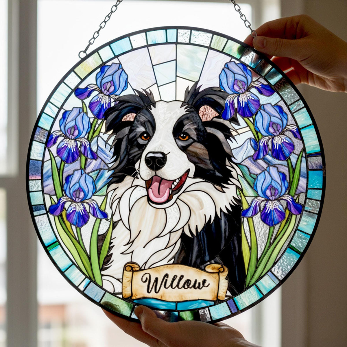 A round stained glass panel featuring a Border Collie dog with blue iris flowers. The dog is black and white with a pink tongue. The name "Willow" is written on a banner at the bottom. The panel is held by hands.