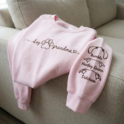 A light pink crewneck sweatshirt featuring dark brown embroidered text that reads dog grandma with a paw print and heart. The sleeve displays three dark brown embroidered dog ear outlines with the names Bumble, Harley Quinn, and Dutchess.