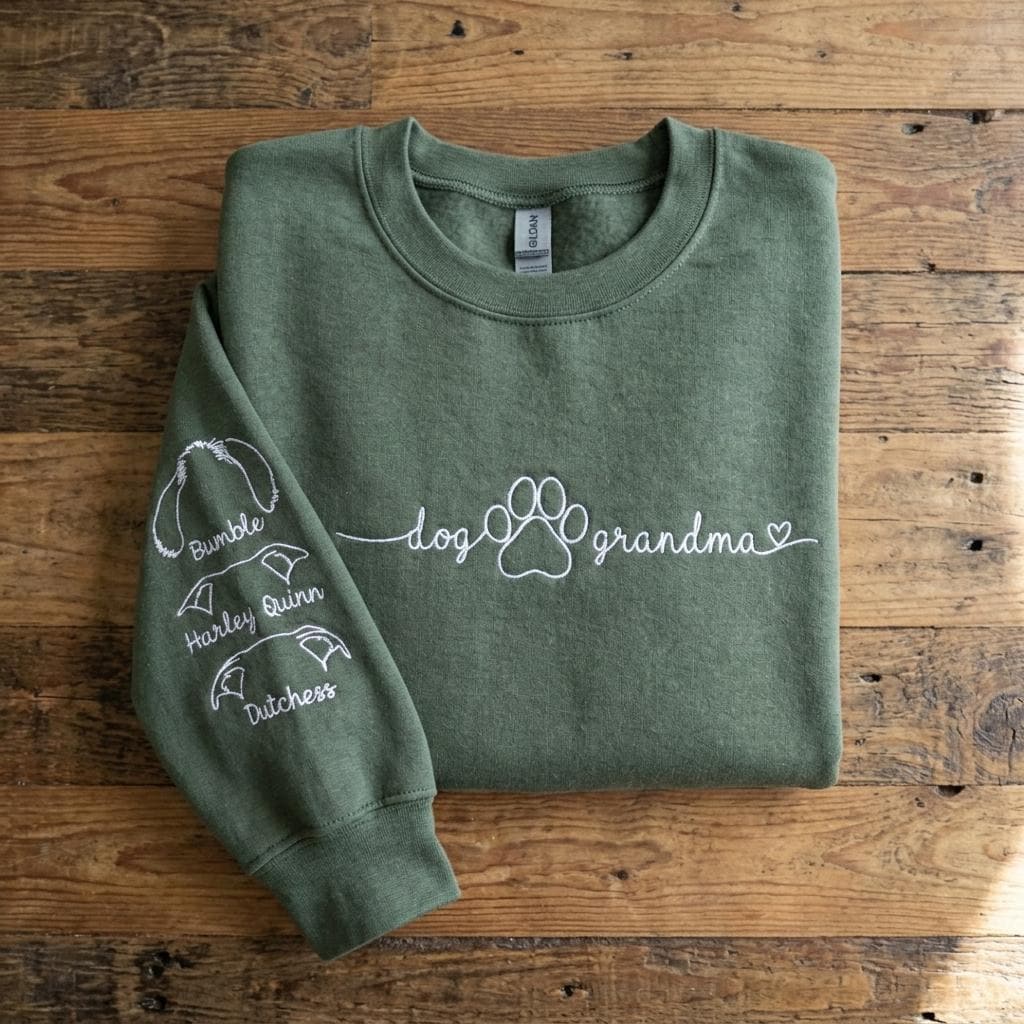 An olive green crewneck sweatshirt featuring white embroidery on the chest that reads dog grandma with a paw print and a heart. The left sleeve includes three white embroidered dog ear outlines with the names Bumble, Harley, and Dutchess.