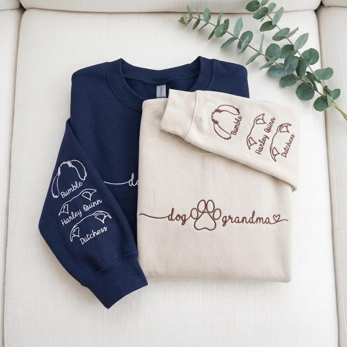 A navy blue sweatshirt and a cream sweatshirt. The navy sleeve features white embroidered dog ear outlines with the names Bumble, Harley Quinn, and Dutchess. The cream sweatshirt features brown text reading dog grandma with a paw print.
