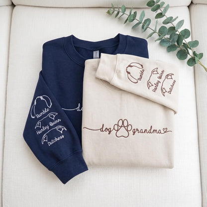 A navy blue sweatshirt and a cream sweatshirt. The navy sleeve features white embroidered dog ear outlines with the names Bumble, Harley Quinn, and Dutchess. The cream sweatshirt features brown text reading dog grandma with a paw print.