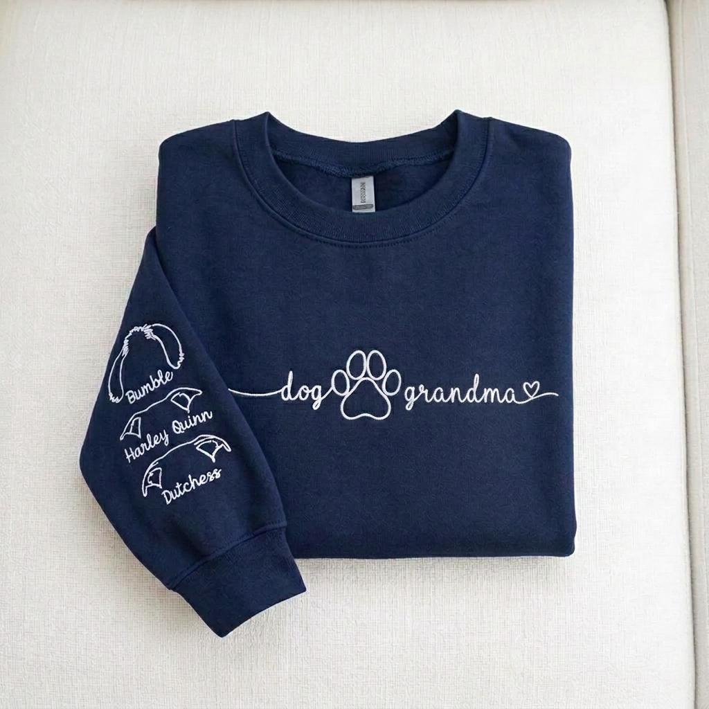 A navy blue crewneck sweatshirt featuring white text on the chest that reads dog grandma with a paw print and heart. The left sleeve displays three white line-art dog ear illustrations with the names Bumble, Harley Quinn, and Dutchess.