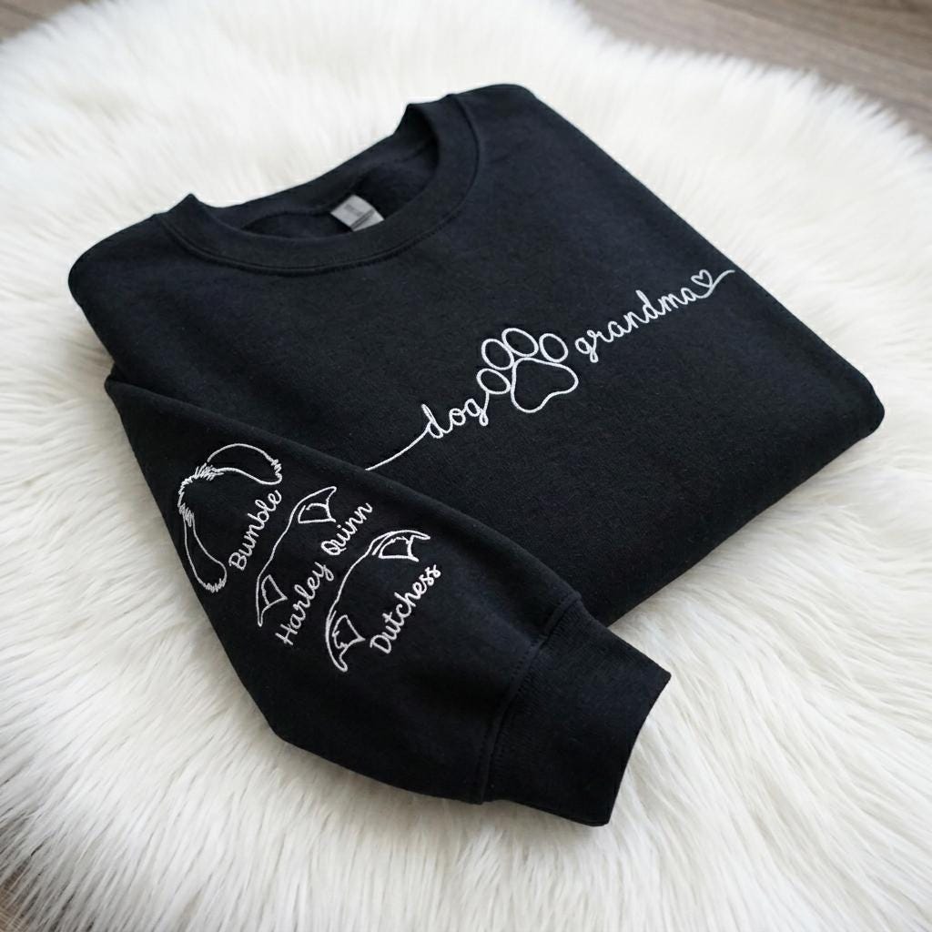 A black long-sleeve sweatshirt featuring white text and graphics. The chest displays the words dog grandma with a paw print and heart. The sleeve features three dog ear outlines with the names Bumble, Harley Quinn, and Dutchess.
