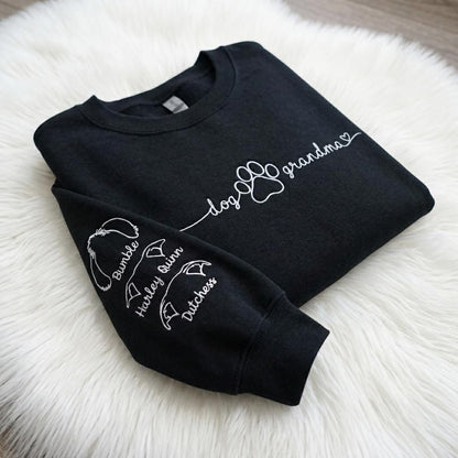 A black long-sleeve sweatshirt featuring white text and graphics. The chest displays the words dog grandma with a paw print and heart. The sleeve features three dog ear outlines with the names Bumble, Harley Quinn, and Dutchess.