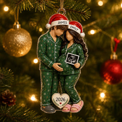 A Christmas ornament featuring a couple in matching green snowflake pajamas and Santa hats labeled James and Emily. The figure on the right holds an ultrasound image. A heart-shaped tag at the bottom displays the year 2026.