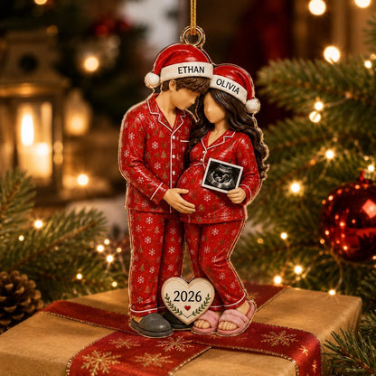 A hanging ornament featuring two figures in red patterned pajamas and Santa hats labeled ETHAN and OLIVIA. The figures hold an ultrasound image, and a heart-shaped plaque at their feet displays the year 2026 in black text.