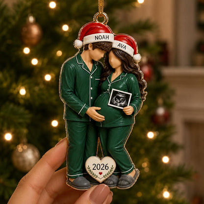 A hanging Christmas ornament featuring two figures in green pajamas and Santa hats labeled NOAH and AVA. The figures stand together, one holding an ultrasound image, with a heart-shaped tag at the bottom displaying the year 2026.