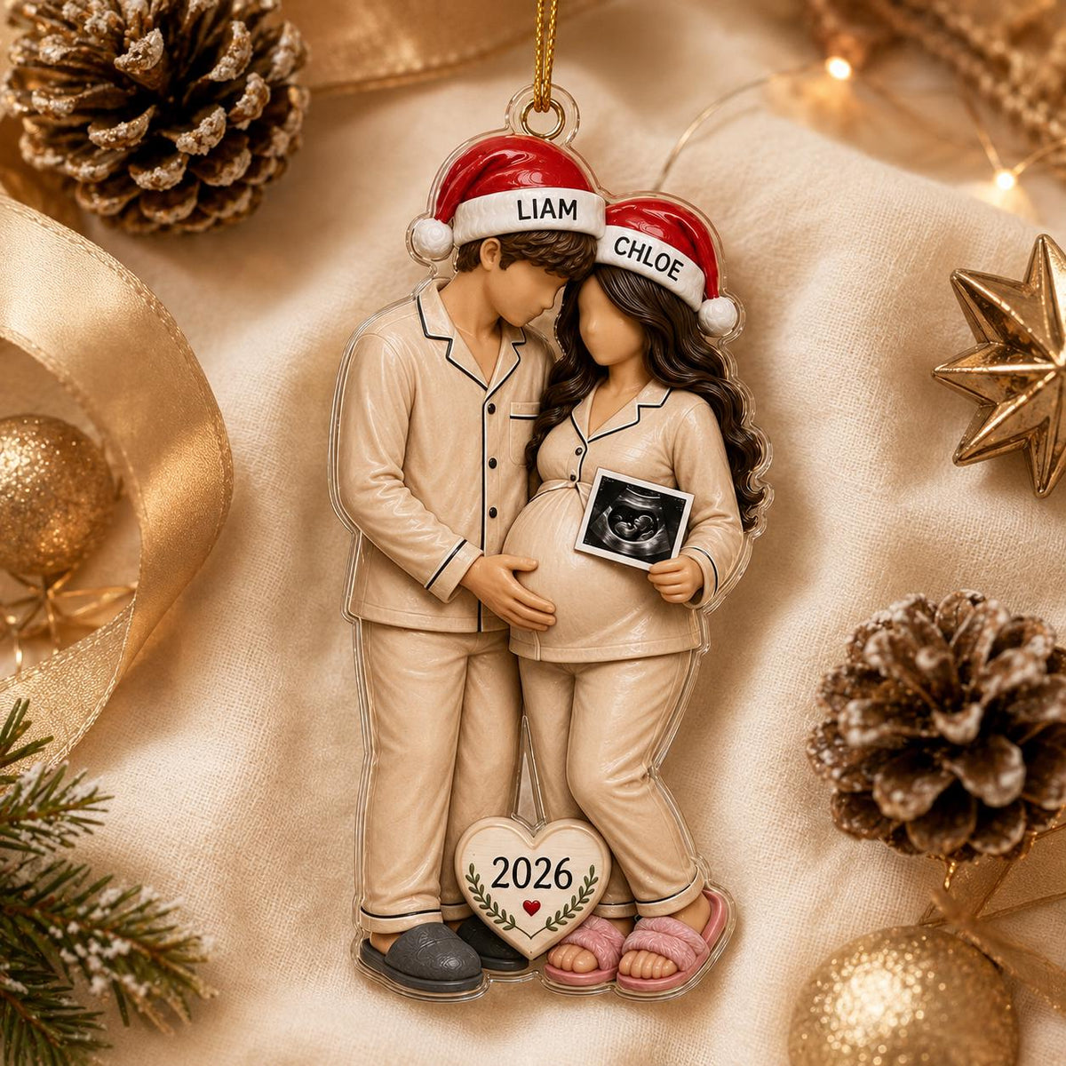 A flat acrylic Christmas ornament featuring a couple in beige pajamas and Santa hats labeled LIAM and CHLOE. The pregnant figure holds a small ultrasound image. A heart-shaped tag at the bottom displays the year 2026 with a red heart.