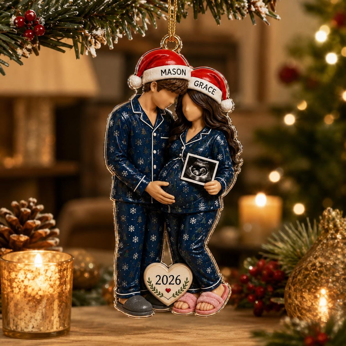 A hanging Christmas ornament featuring two figures in matching navy blue snowflake pajamas and red Santa hats. The hats are labeled MASON and GRACE. The figure on the right holds a sonogram image. A heart at the base displays 2026.