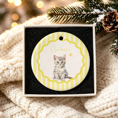 A round ceramic ornament featuring a yellow and white striped background with a scalloped border. The center displays a realistic illustration of a grey tabby kitten and the name Cookie written in cursive, accompanied by a small paw print.