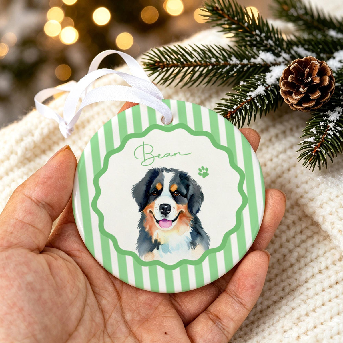 A round ceramic ornament featuring a white center with a watercolor illustration of a Bernese Mountain Dog, the name Bean in script, and a green paw print. The border has a scalloped edge and light green and white vertical stripes.