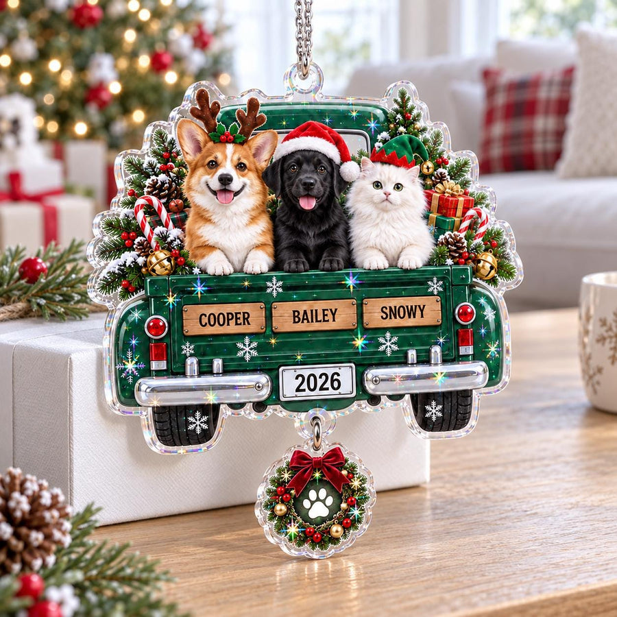 A Christmas ornament shaped like a green pickup truck carrying a Corgi, a black Labrador, and a white cat. The truck features the names COOPER, BAILEY, and SNOWY, the year 2026, and a dangling wreath charm with a white paw print design.