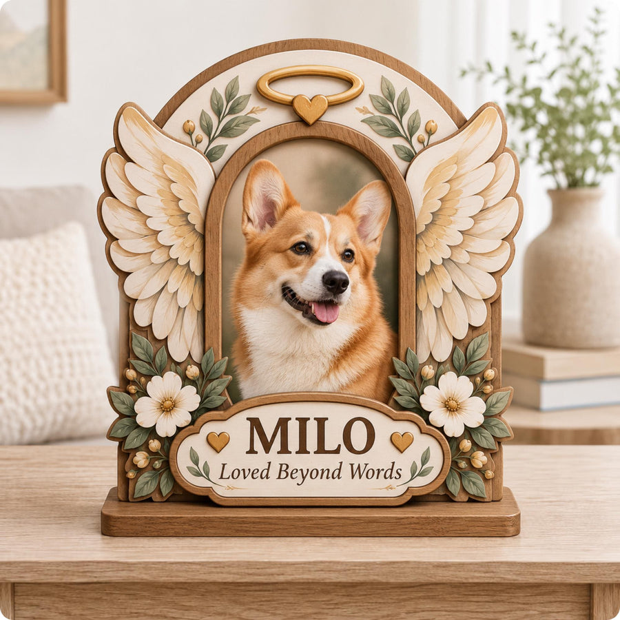 A wooden memorial photo frame featuring a portrait of a corgi. The frame is decorated with sculpted angel wings, white flowers, green leaves, and a gold halo with a heart. A plaque reads MILO and Loved Beyond Words with two hearts.