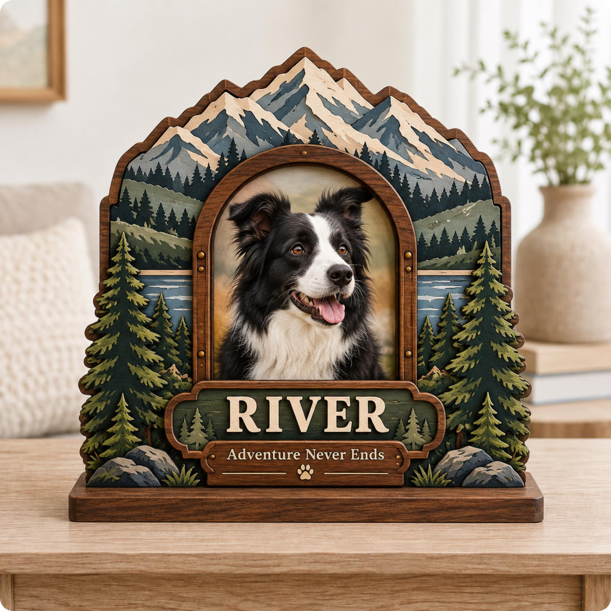 A wooden picture frame featuring a layered mountain and forest landscape with a central portrait of a black and white dog. The frame includes a plaque with the name RIVER and the text Adventure Never Ends with a paw print icon.