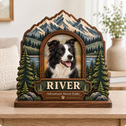 A wooden picture frame featuring a layered mountain and forest landscape with a central portrait of a black and white dog. The frame includes a plaque with the name RIVER and the text Adventure Never Ends with a paw print icon.