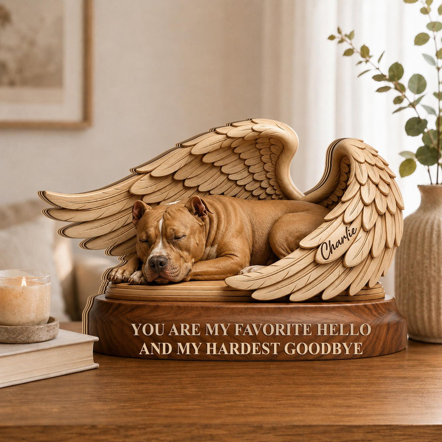 A wooden memorial sculpture featuring a brown dog sleeping peacefully between two large, detailed angel wings. The base is engraved with the text, "YOU ARE MY FAVORITE HELLO AND MY HARDEST GOODBYE," and the name "Charlie" is etched on one wing.