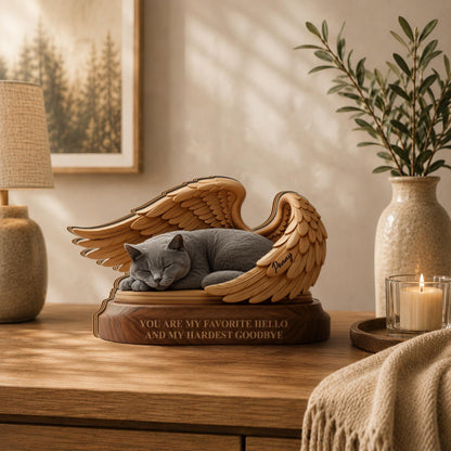 A wooden memorial sculpture featuring a grey cat sleeping inside a pair of carved angel wings. The base is engraved with the text, "YOU ARE MY FAVORITE HELLO AND MY HARDEST GOODBYE," and the name "Penny" is etched on the wing.