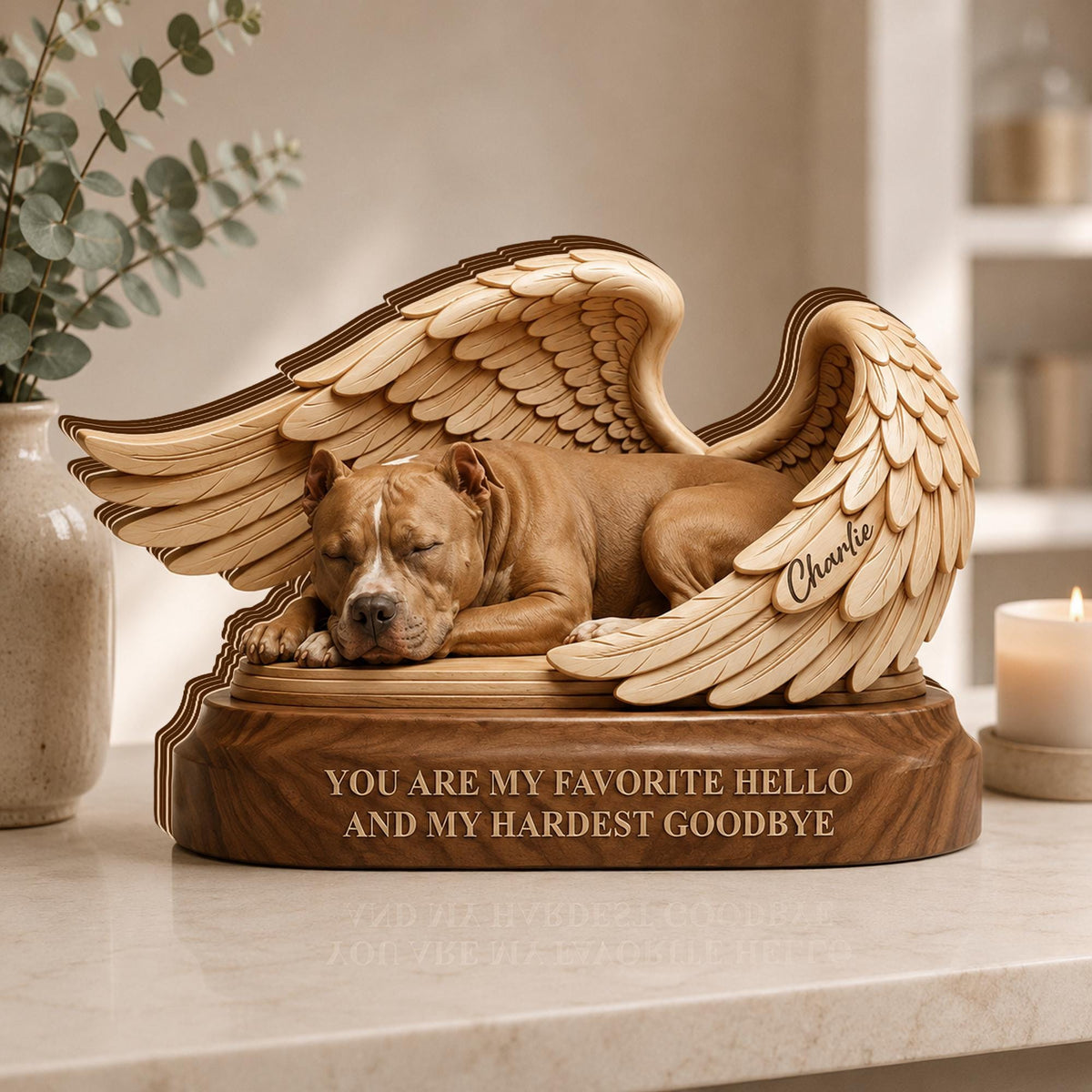 A wooden memorial sculpture featuring a sleeping brown pit bull dog nestled between two large, detailed carved wings. The base is engraved with the text, "YOU ARE MY FAVORITE HELLO AND MY HARDEST GOODBYE," and the name "Charlie" is etched on one wing.