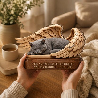 A wooden memorial sculpture featuring a sleeping gray cat nestled between two carved wings. The base is engraved with the text "YOU ARE MY FAVORITE HELLO AND MY HARDEST GOODBYE" and the name "Penny" is etched onto one of the wings.