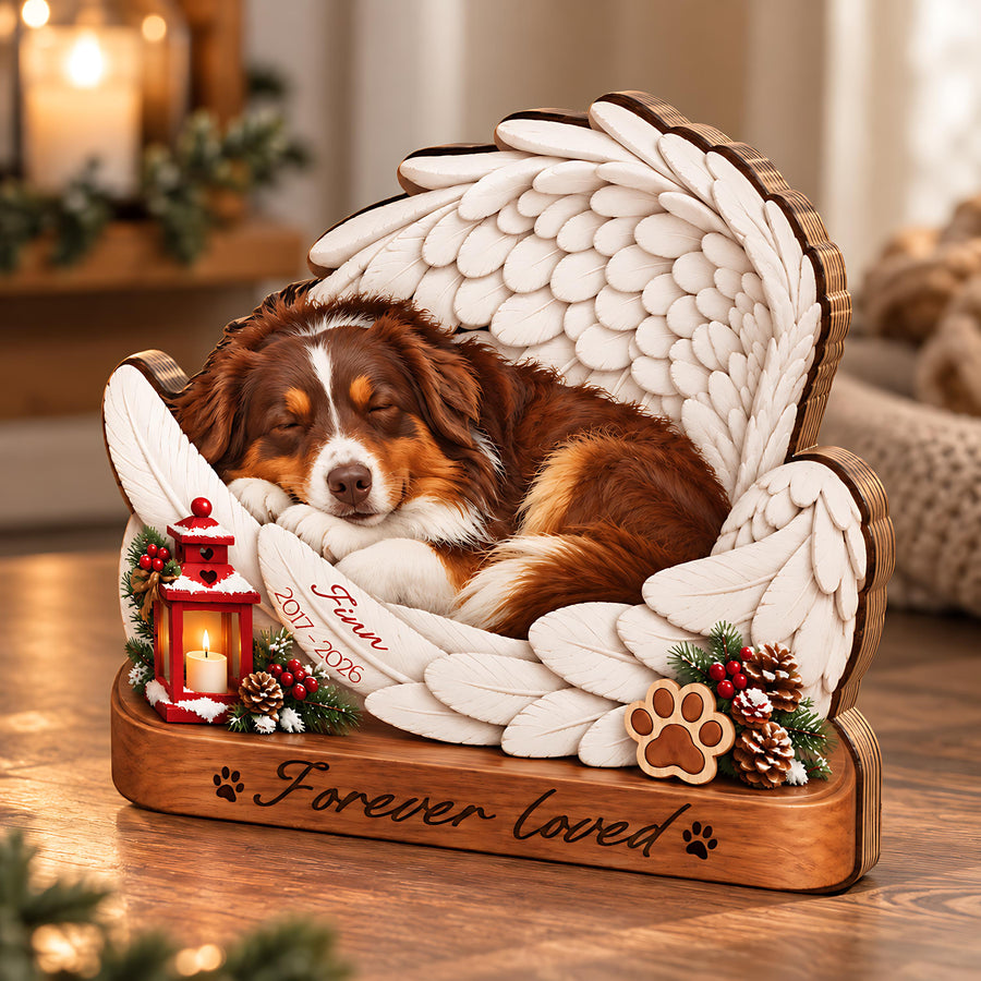 A wooden pet memorial featuring a sleeping brown and white dog nestled within large white angel wings. The base is engraved with "Forever loved" and paw prints. A red lantern, pinecones, and a feather inscribed "Finn 2017-2026" are included.