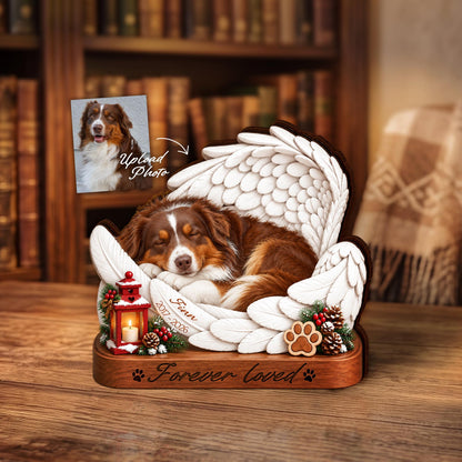 A wooden memorial plaque featuring a sleeping brown and white dog nestled within white angel wings. The base includes a red lantern, pinecones, a paw print, and the text "Forever loved" with "Finn 2017-2026" on the wing.