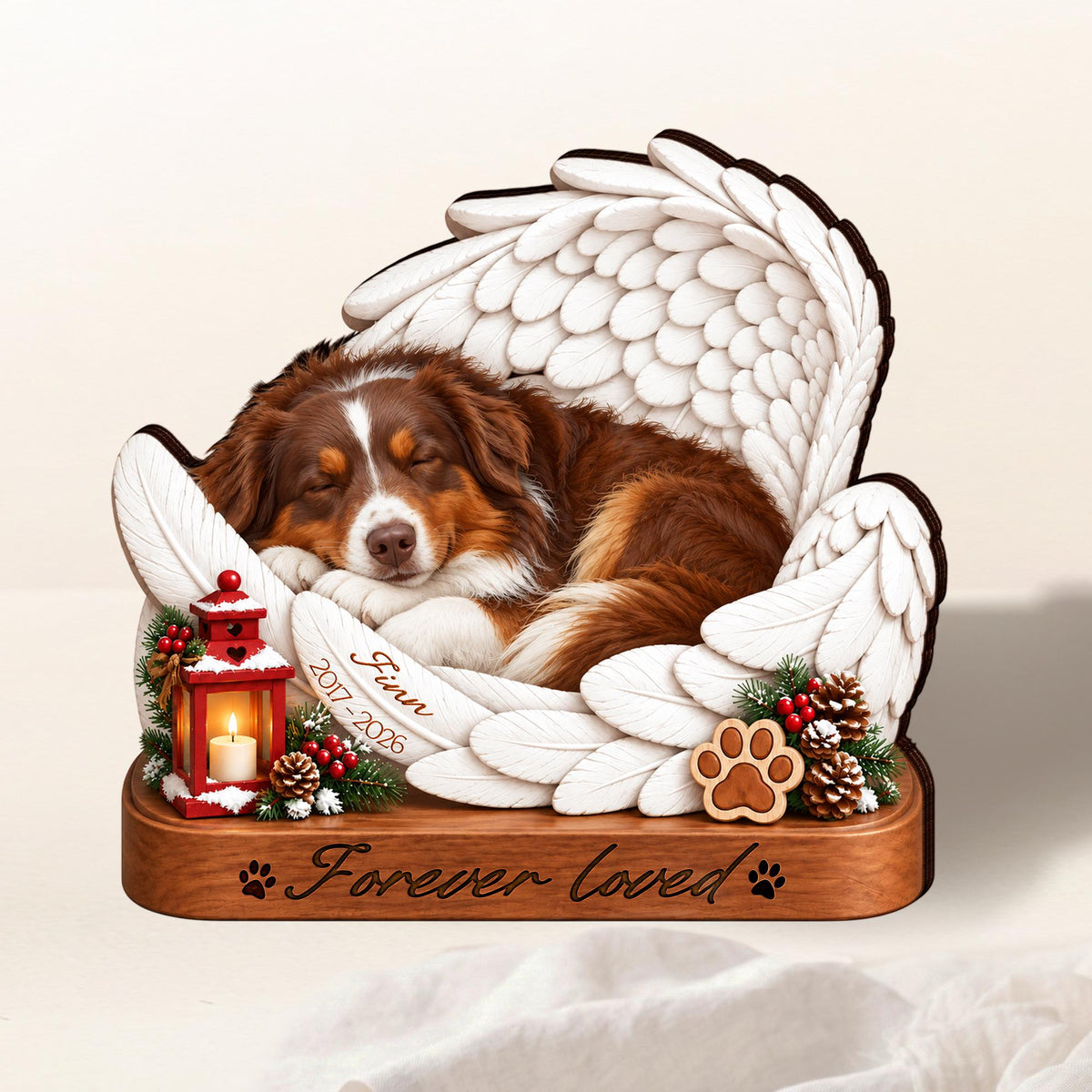 A memorial ornament featuring a brown and white dog sleeping inside large white angel wings. A red lantern with a lit candle, pine cones, and berries sit on a wooden base engraved with "Forever loved" and paw prints. The name "Finn 2017-2026" is inscribed on a feather.